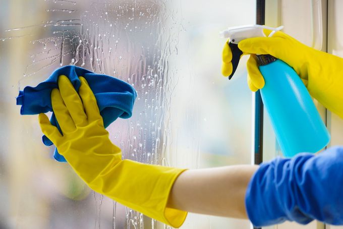 Gloved hands cleaning a foggy window with a blue cloth and spray bottle