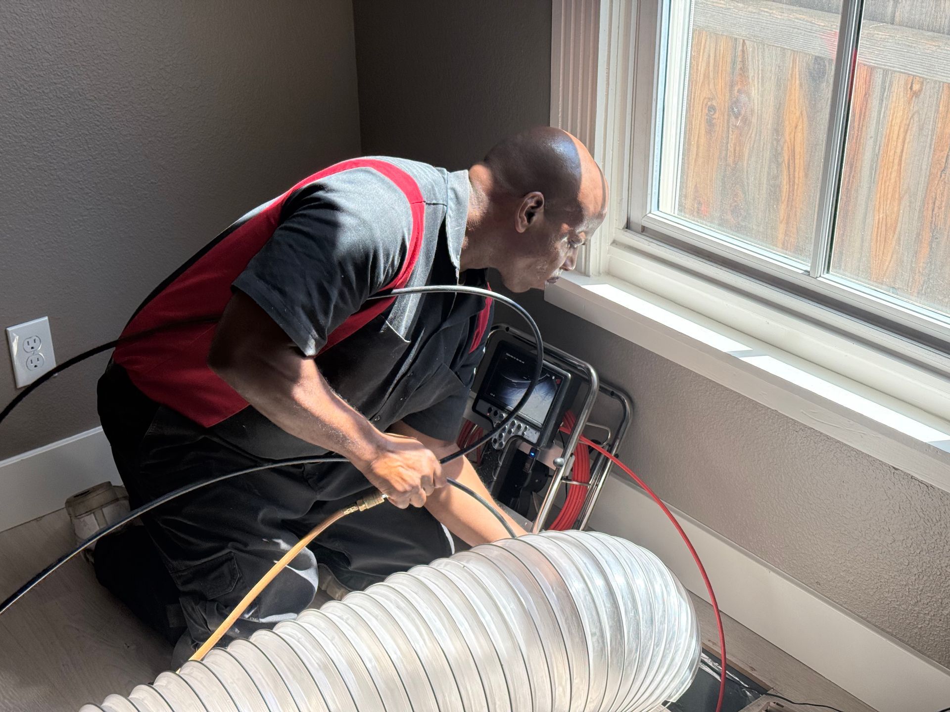 Man inspecting with camera equipment near a window. He is wearing a red and black uniform in a room with a white vent.