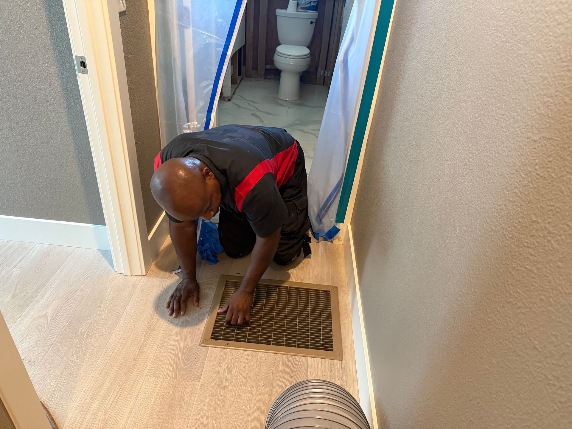 Man kneeling, cleaning floor vent in a bathroom. A plastic sheet covers the doorway.