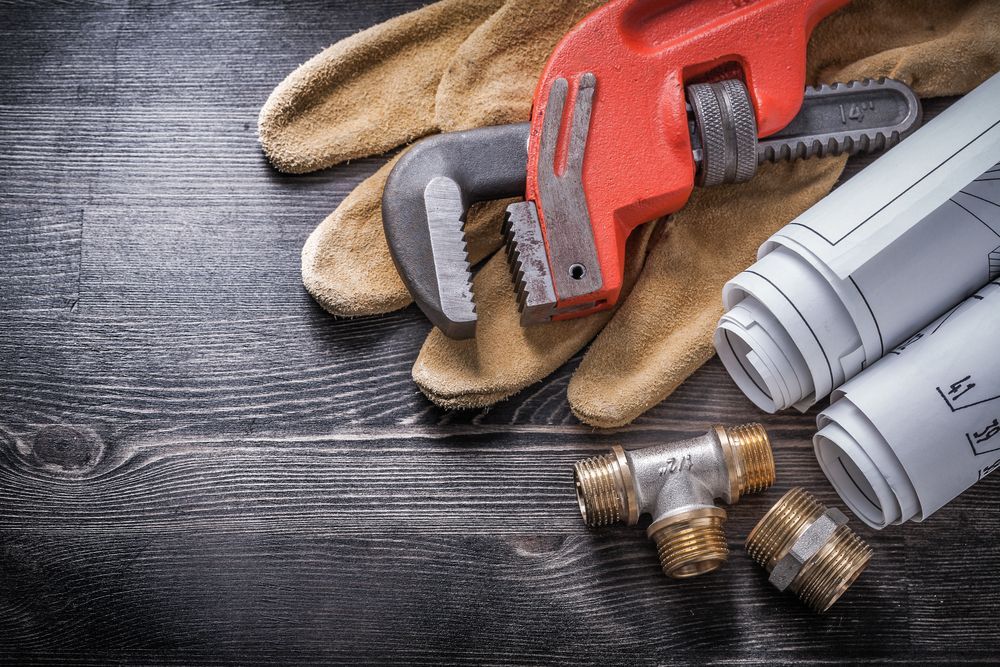 A Pair of Plumbing Gloves, a Wrench, Pipes, and Blueprints on a Wooden Table — Subat Plumbing Services in Kurri Kurri, NSW
