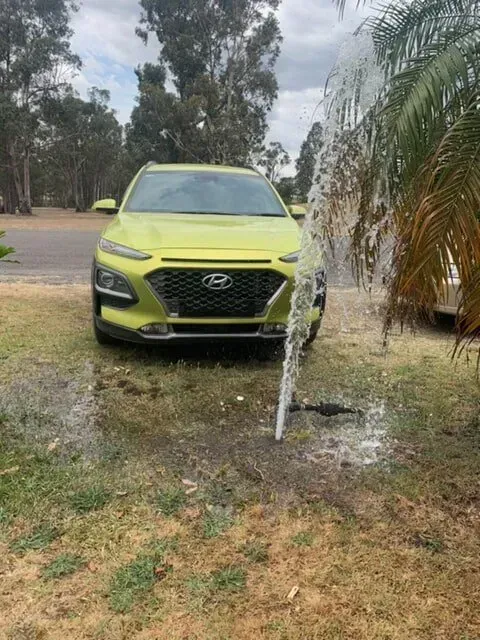 A Yellow Hyundai Kona is Parked in Front of a Hose Spraying Water — Subat Plumbing Services in Kurri Kurri, NSW