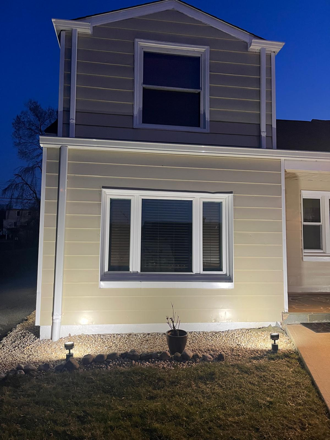 Two-story beige house at dusk, lit by exterior lights, with white-trimmed windows and a small front lawn.