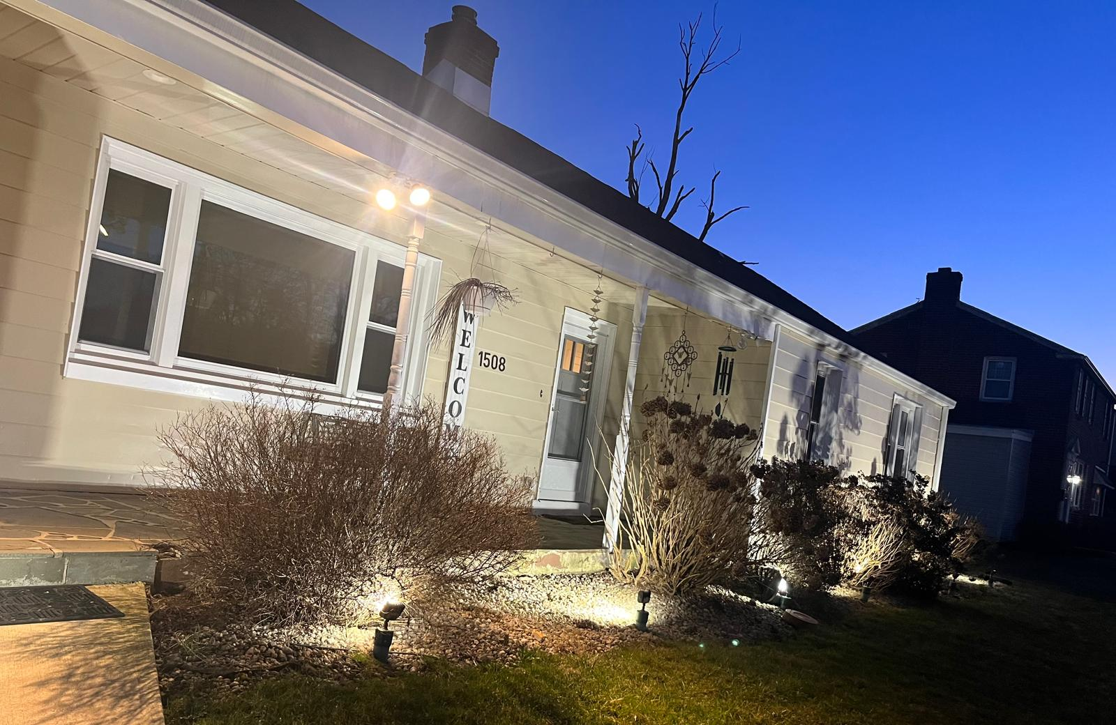 Single-story house at dusk with lit windows, shrubs, and a front walkway under a blue sky.