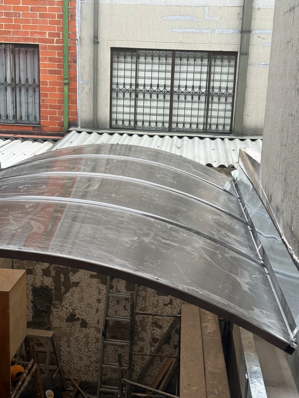 Un toldo curvo con paneles transparentes cubre un área exterior, con ventanas y un edificio de ladrillo visibles en el fondo.