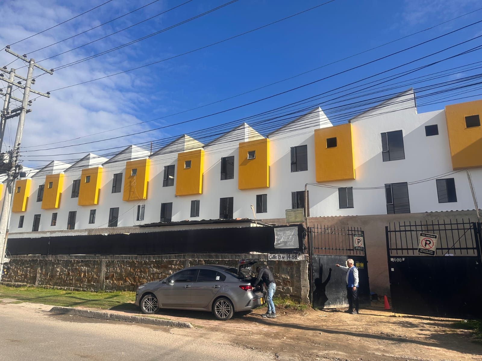Hilera de casas blancas con detalles en amarillo; gente cerca de un coche y una puerta.
