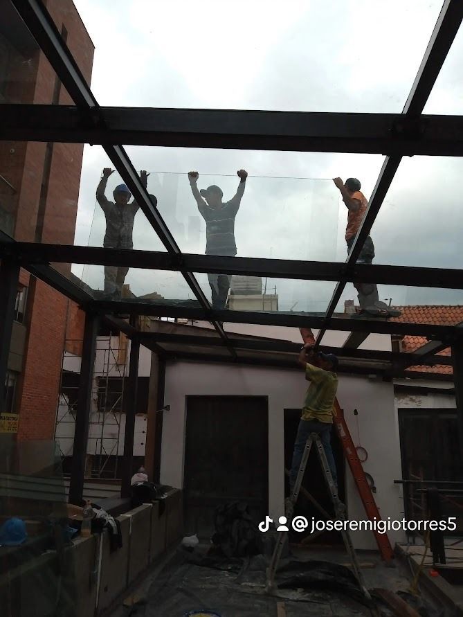 Trabajadores instalando paneles de vidrio en una estructura de techo con marco de metal, cielo nublado.