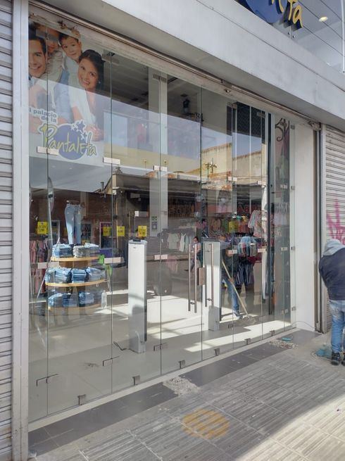 Tienda con puertas de cristal. Vaqueros expuestos en el interior, un póster con gente en el escaparate. Persianas metálicas cerradas a izquierda y derecha.