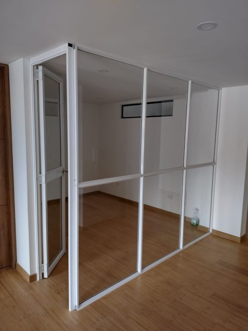 Mampara de vidrio con marco blanco y puerta en habitación con suelo de madera y paredes blancas.