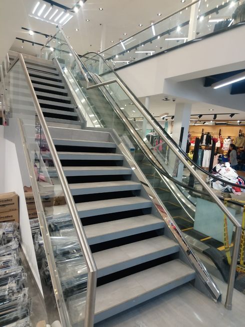 Escalera y escalera mecánica en un centro comercial. Barandillas de vidrio y acero, escalones grises y clientes al fondo.