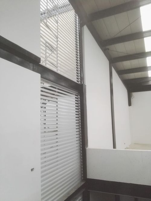 Vista interior de un edificio con paredes blancas y una ventana de vidrio corrugado y vigas de metal.