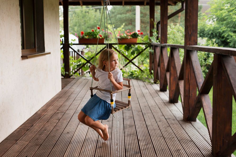 Menina balançando em um balanço de varanda, deck de madeira com corrimão e flores ao fundo.
