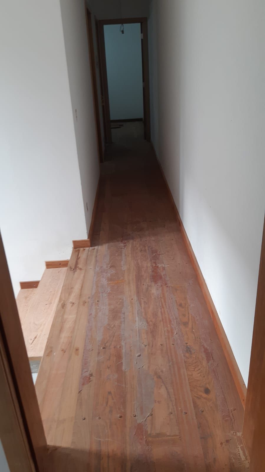 Escada e corredor de madeira, que levam a uma porta. Paredes brancas, piso de madeira.