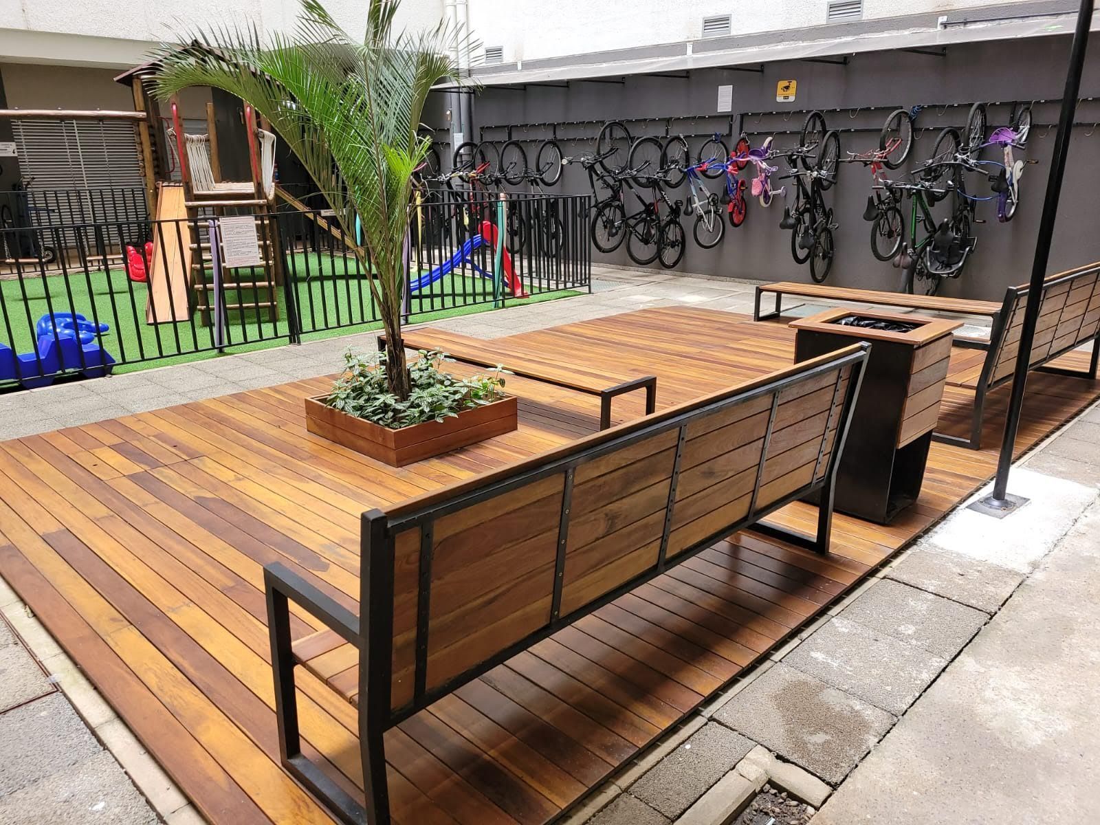Deck de madeira com assentos, suporte para bicicletas, pequeno parque infantil e uma palmeira em uma área externa.