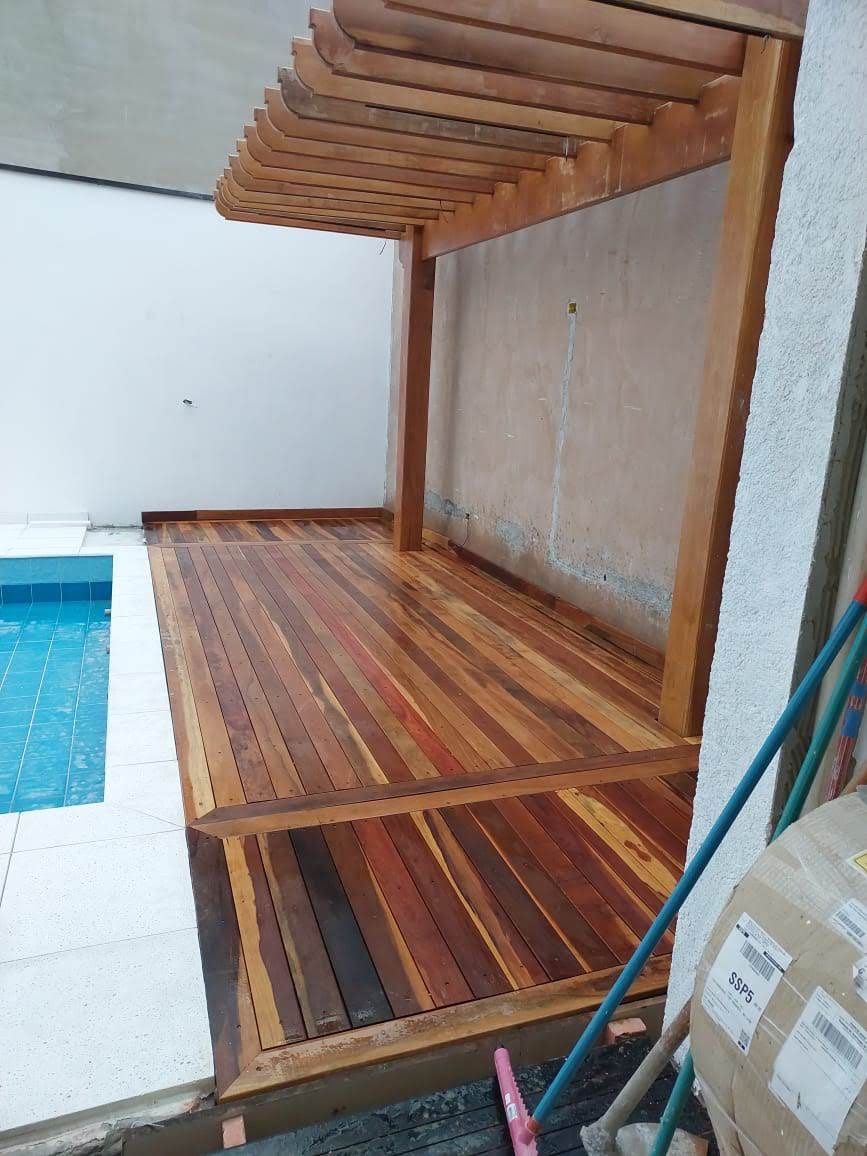 Deck de madeira e pérgola ao lado da piscina. Madeira marrom, água azul, paredes brancas.