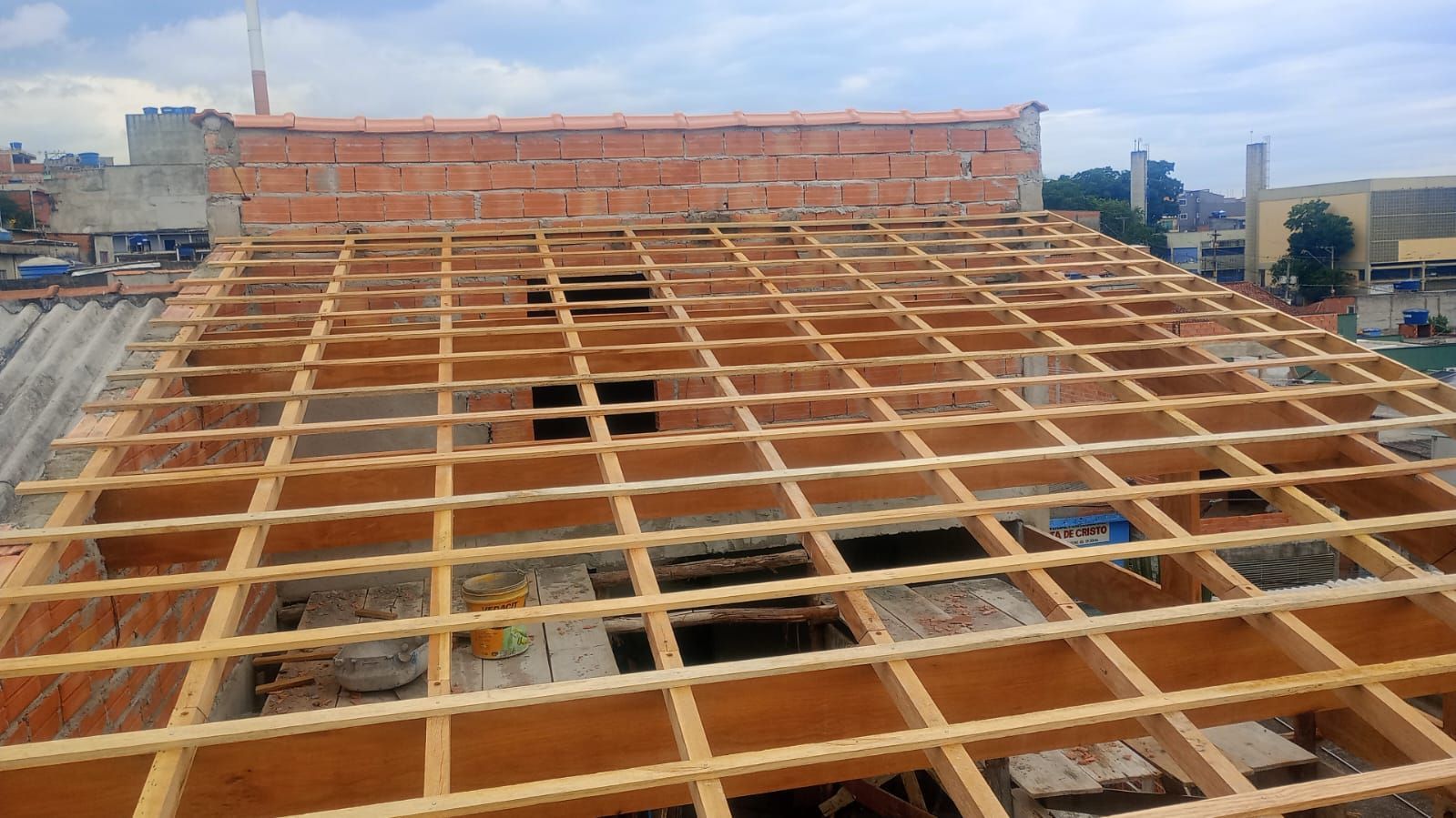 Estrutura de madeira de um telhado em construção. Parede de tijolos ao fundo.