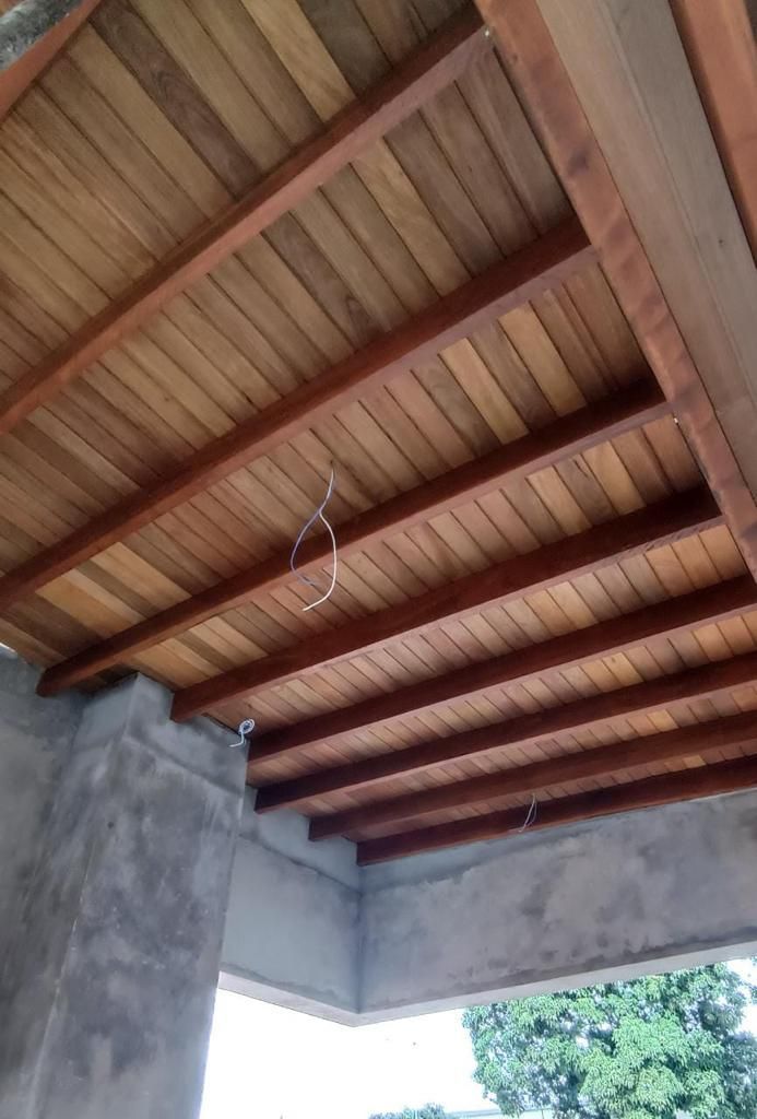 Teto de madeira com vigas escuras e tábuas de madeira clara, visto de baixo. Paredes de concreto cinza.