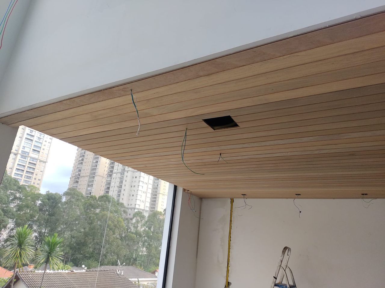 Teto com painéis de madeira clara, caixa de energia elétrica e fiação, cobrindo parcialmente uma janela com vista para a cidade.