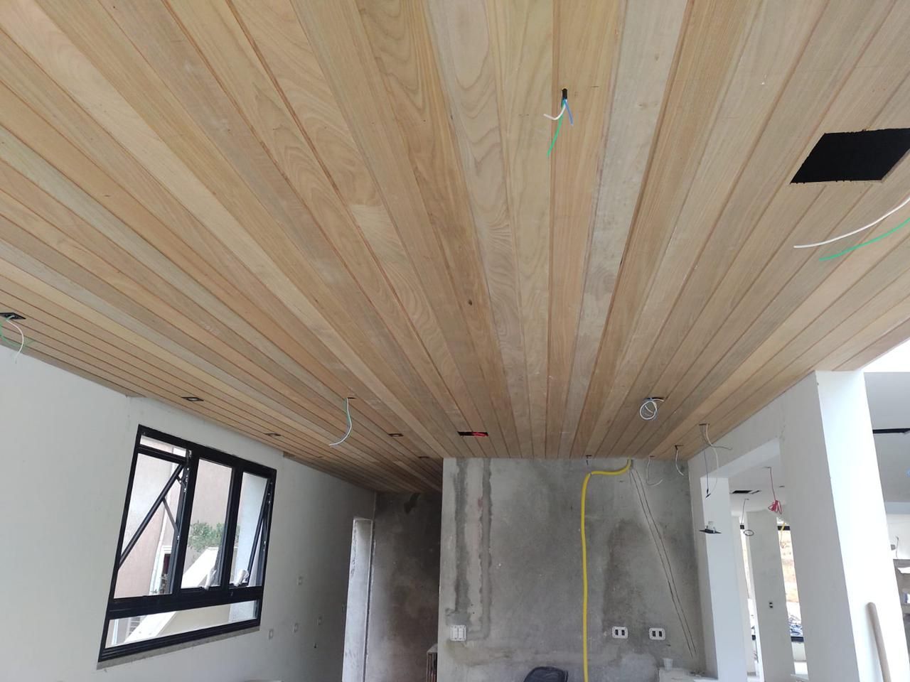 Instalação de forro de madeira em um cômodo em construção com fiação elétrica e janela preta.