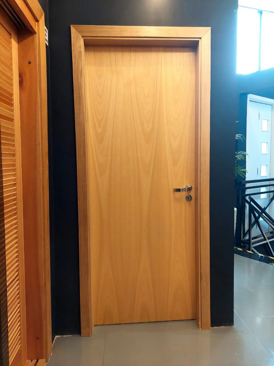 Porta interna de madeira com moldura marrom-clara, puxador e parede preta ao fundo.