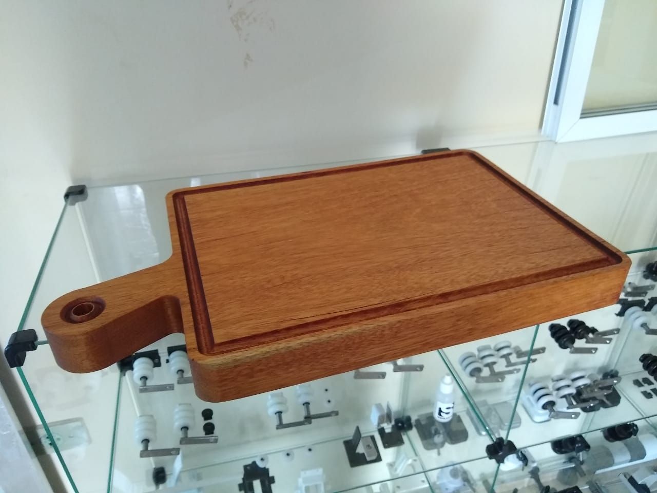 Tábua de corte retangular de madeira com alça, sobre uma prateleira de vidro.