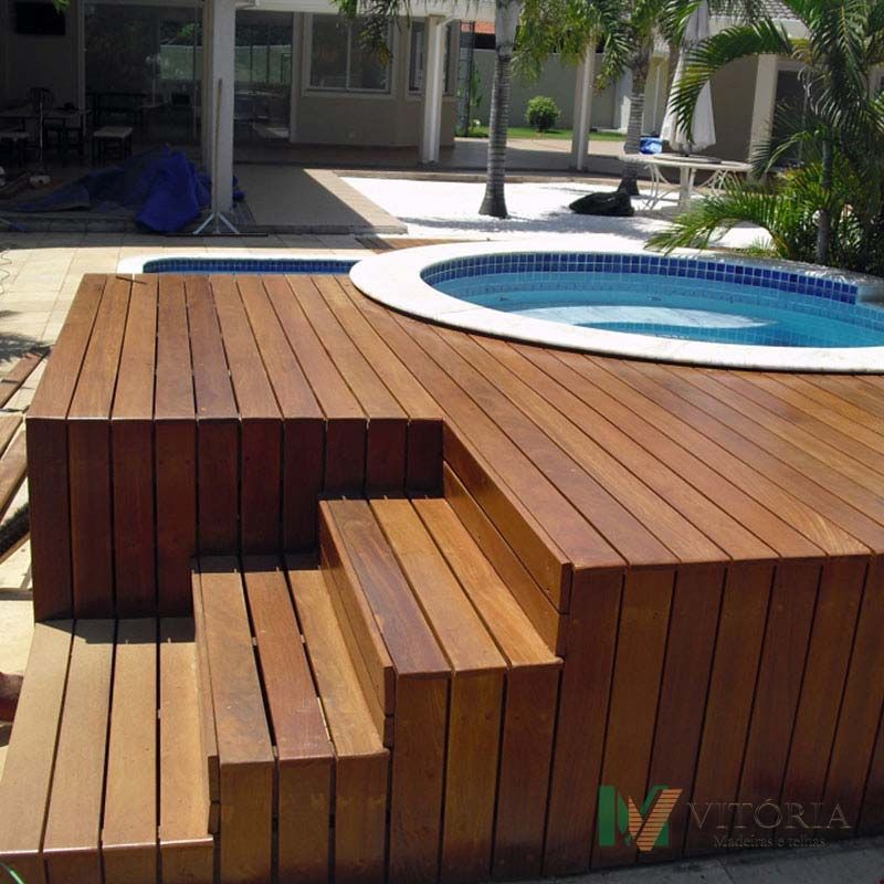 Um deck de madeira com degraus circunda uma jacuzzi circular. Ao fundo, vê-se uma pequena piscina e uma casa.