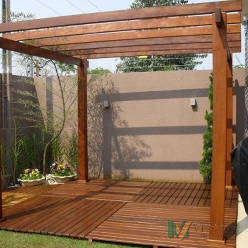Pérgola de madeira sobre um deck, em um quintal. Madeira marrom, parede bege, grama verde.