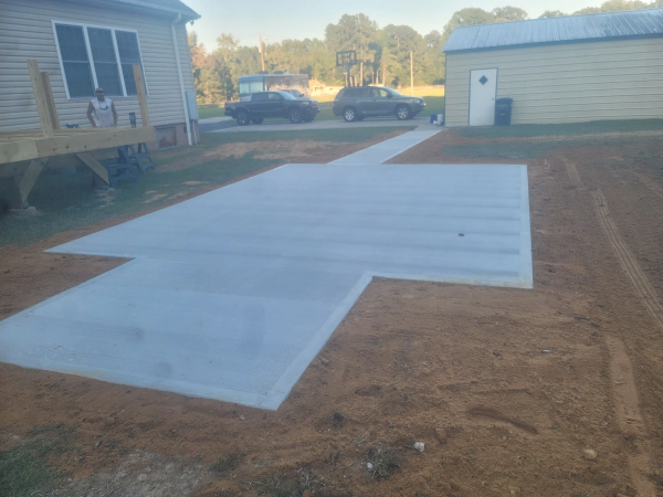Newly poured concrete slab and walkway in a rural yard, with a house and shed in the background.