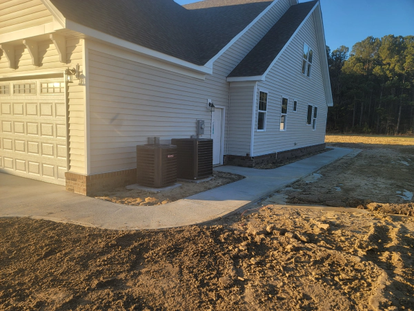 A concrete sidewalk curves past a light-colored house with garage, HVAC units, and bare earth.