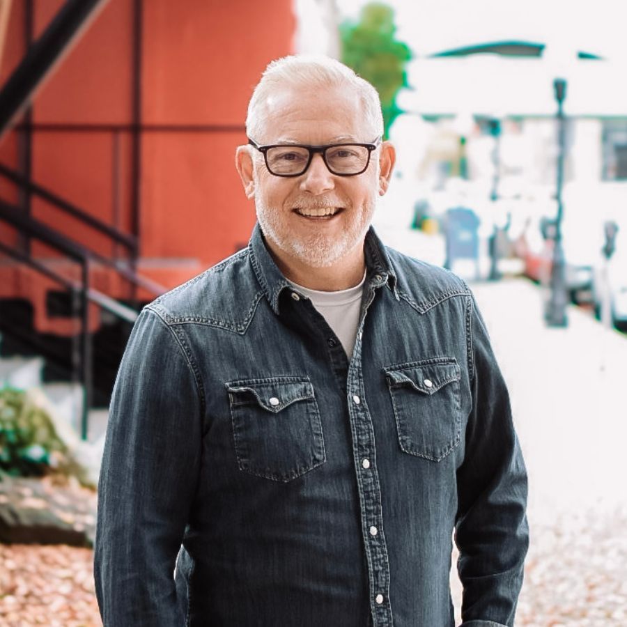 Man with glasses, smiles outdoors, wearing a denim shirt and white undershirt. Red building backdrop.
