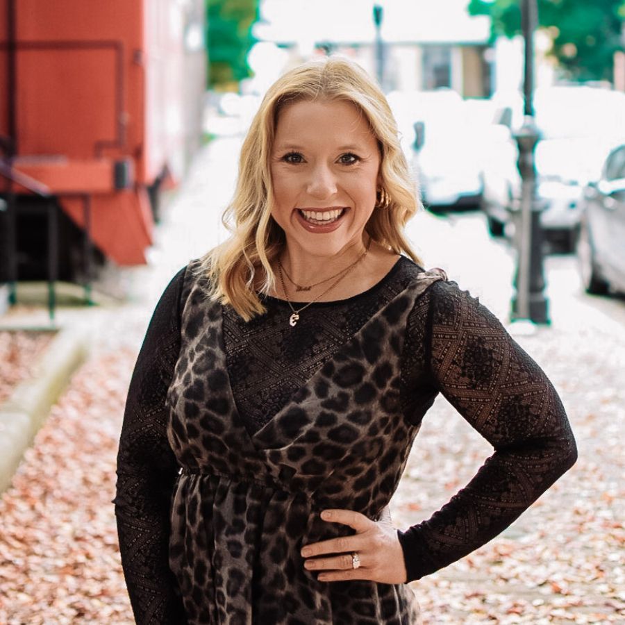Woman smiles, hand on hip, wearing a leopard print dress. Outdoor setting with fall foliage, buildings and parked cars in background.
