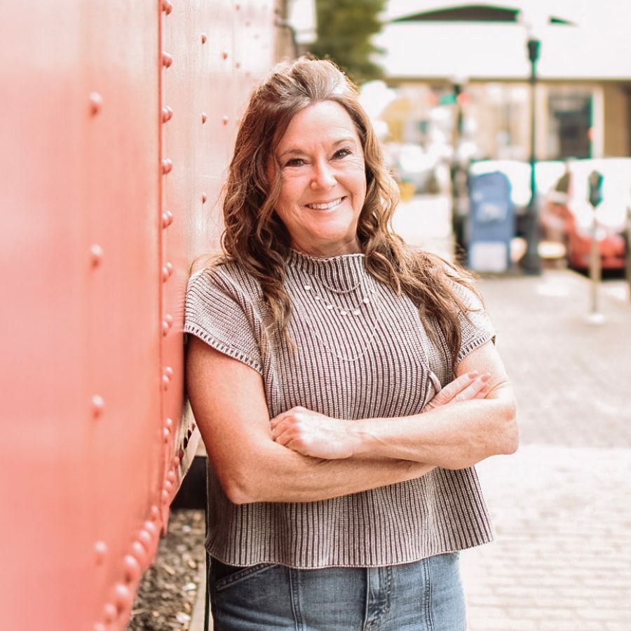 Woman with arms crossed, smiling, leaning against a red train car in a town square.
