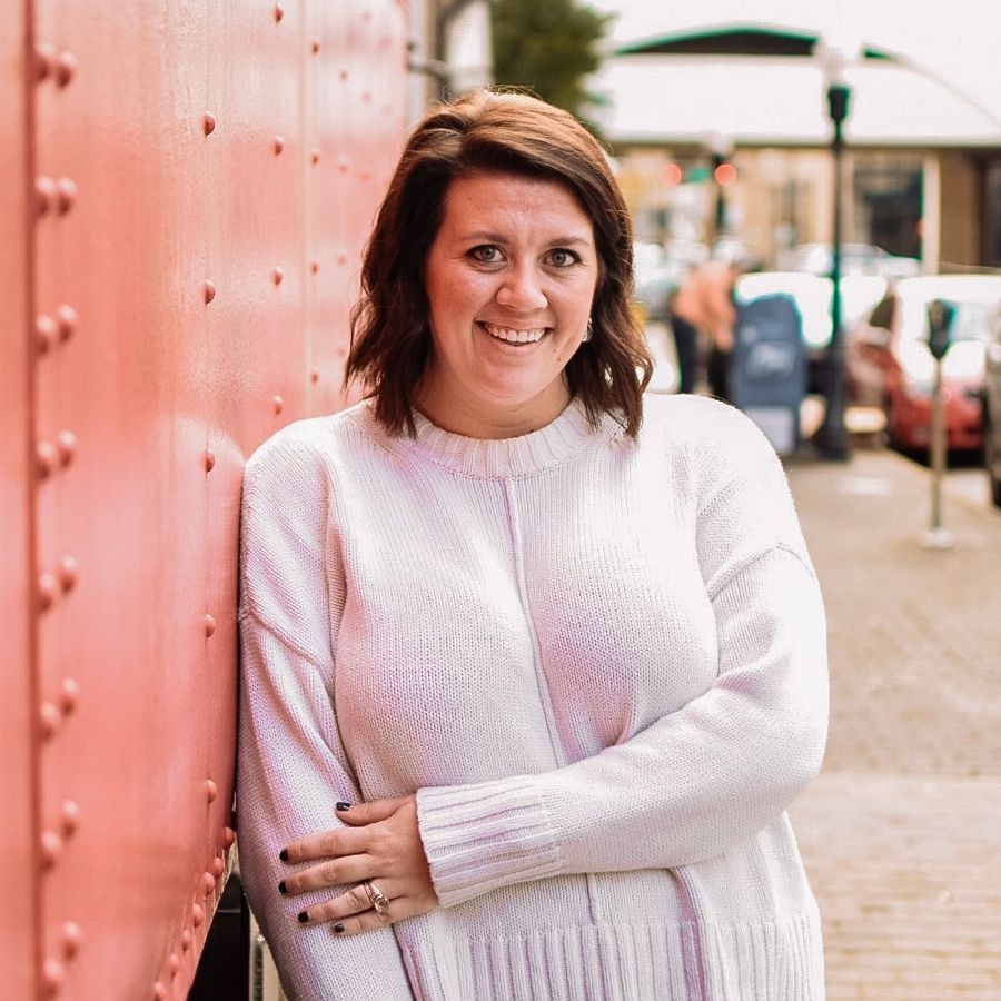 Woman in a white sweater smiles, leaning against a coral wall, in an outdoor urban setting.