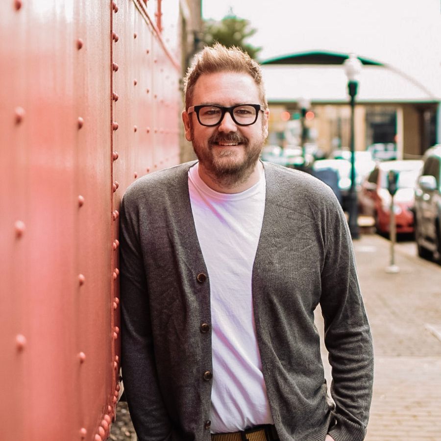 Man in glasses, gray cardigan, white shirt, smiles, leans against a pink wall on a city street.