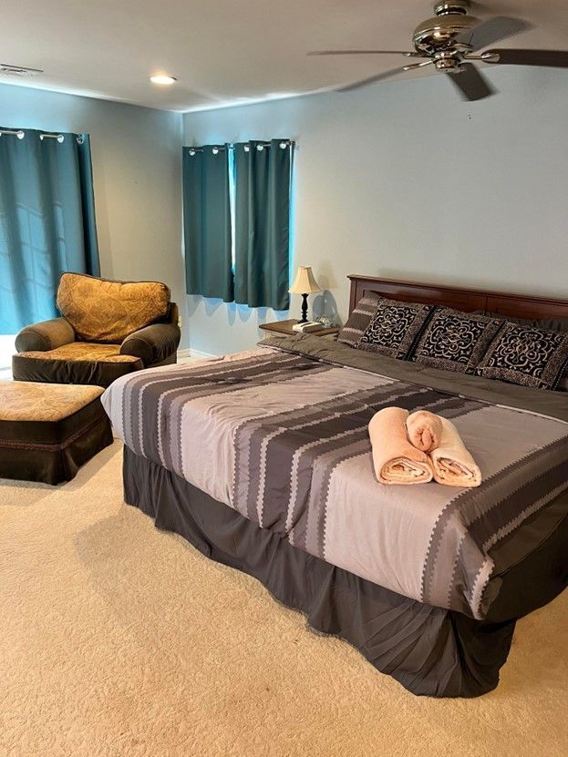 A bedroom with a king size bed , chair , ottoman and ceiling fan.