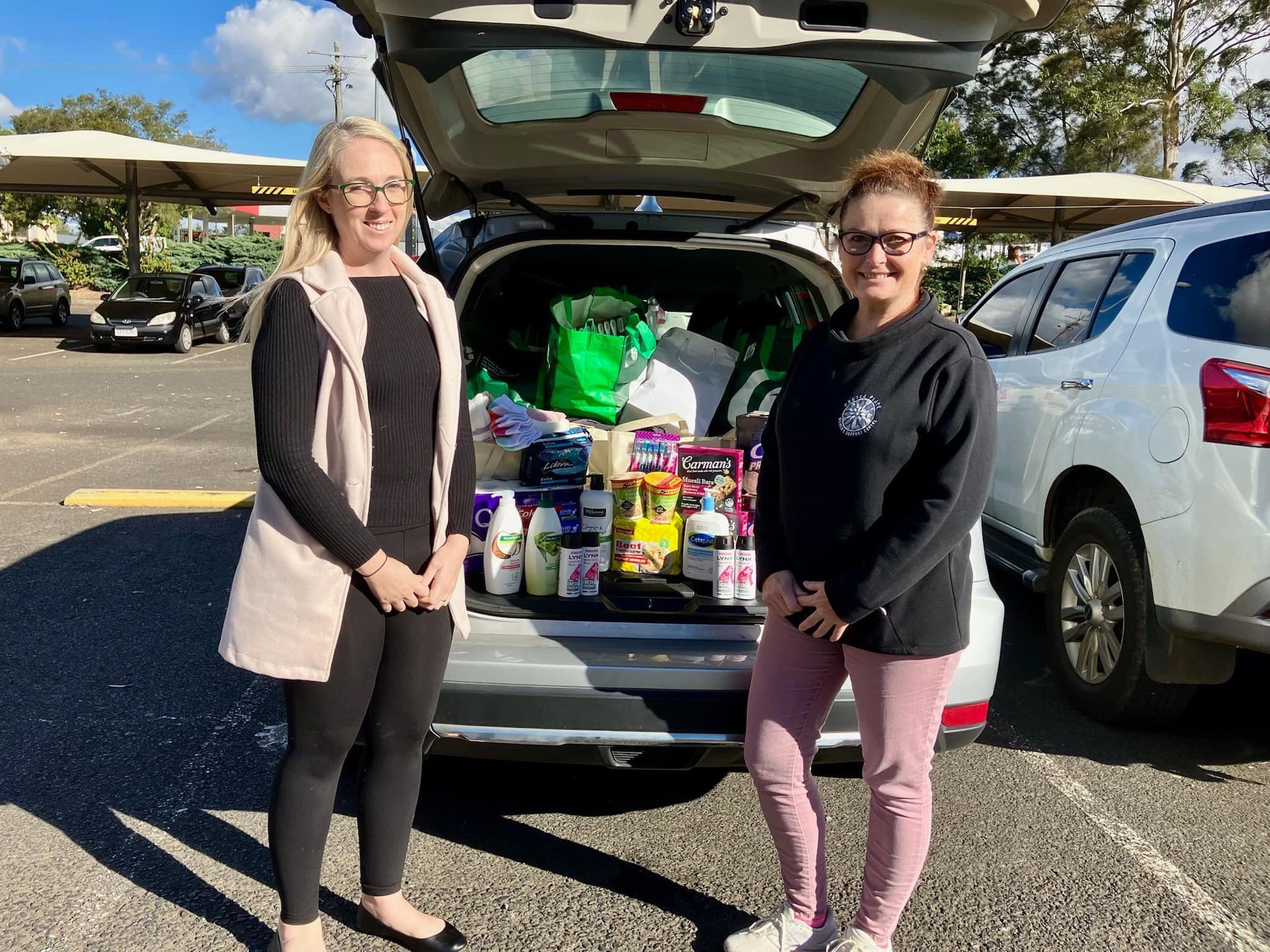 Two women are standing in front of a car filled with groceries.