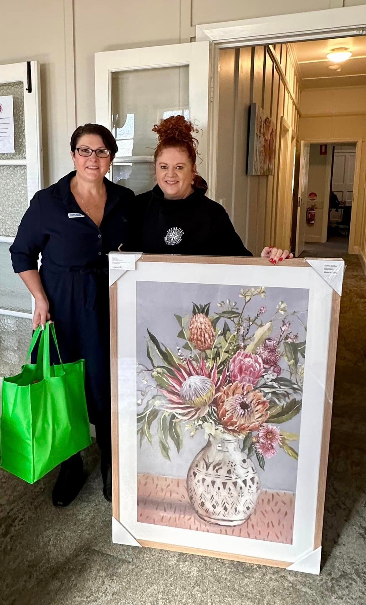 Two women are standing next to each other holding a framed painting of flowers in a vase.