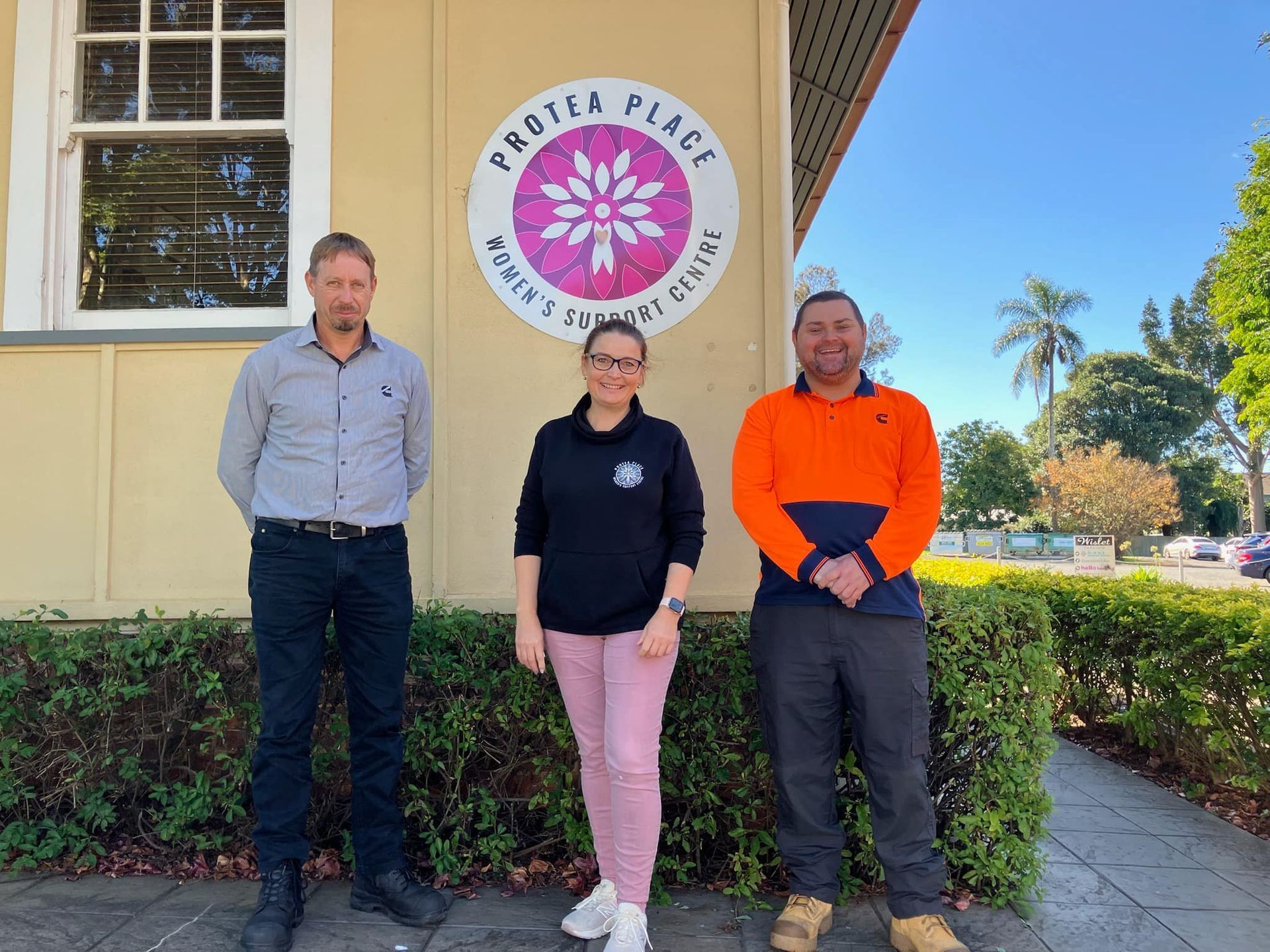 Three people are posing for a picture in front of the Protea Place building.