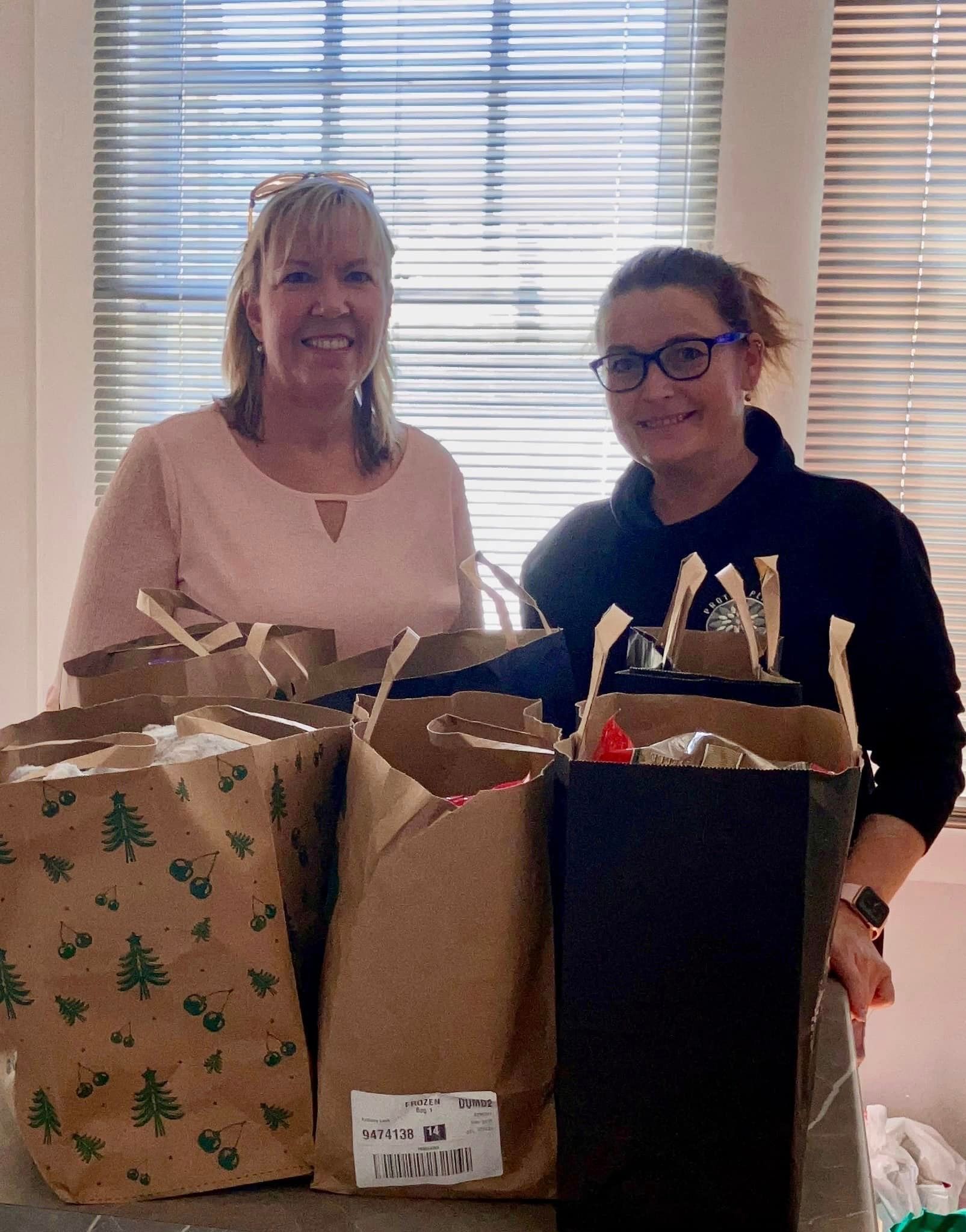 Two women are standing next to each other holding bags of gifts.