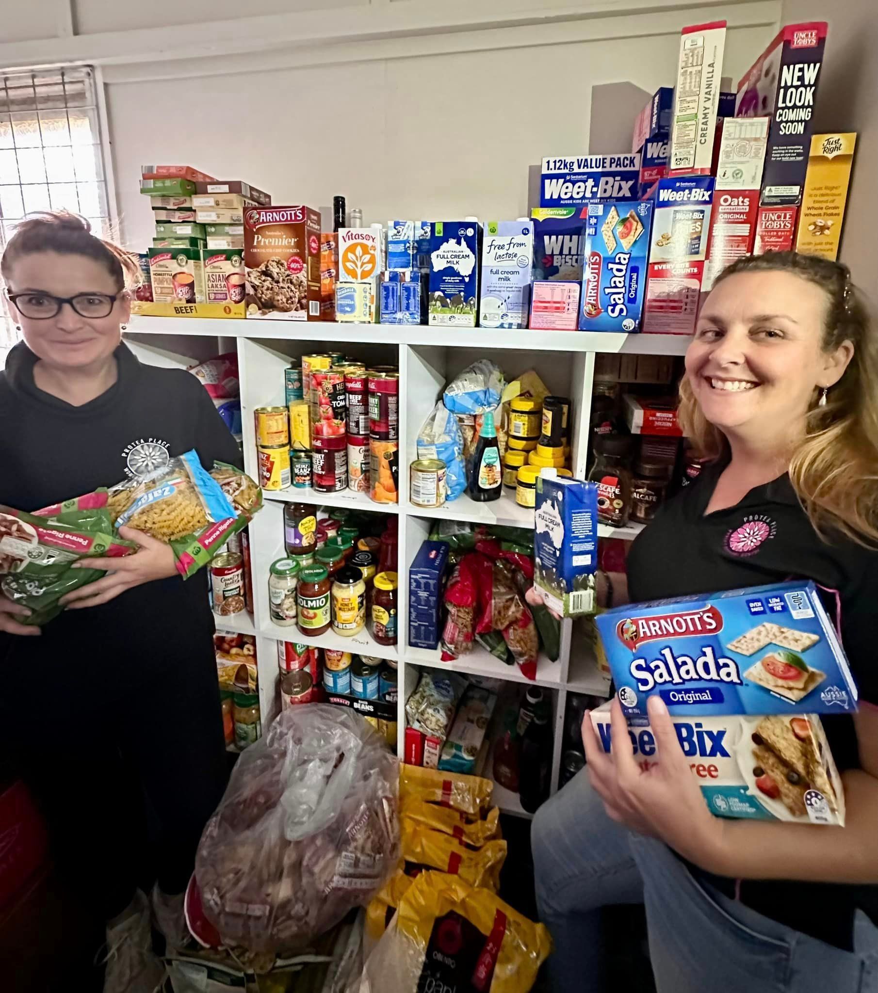 Two women are holding boxes of food in front of a shelf full of food.