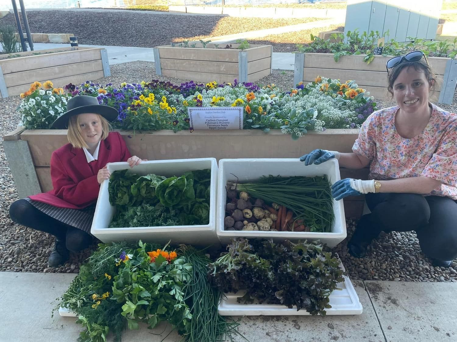 A woman and a child are kneeling next to a planter filled with vegetables.