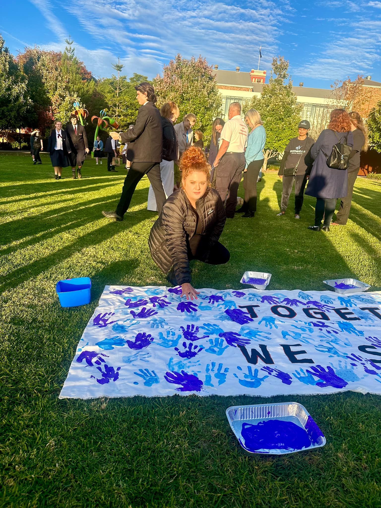 A woman is kneeling on the grass next to a large piece of paper with handprints on it.