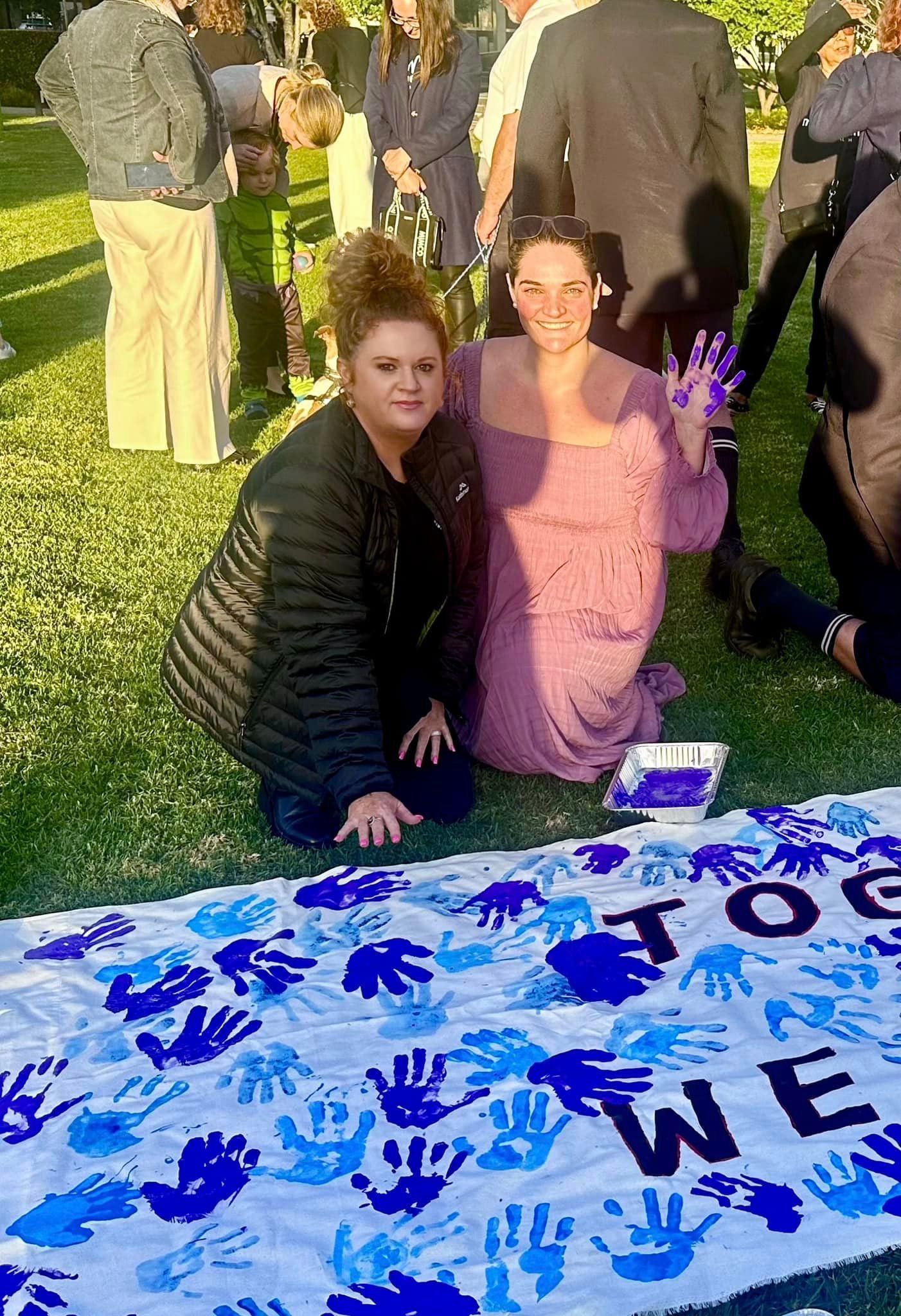 Two women are sitting on the grass next to a blanket with handprints on it.