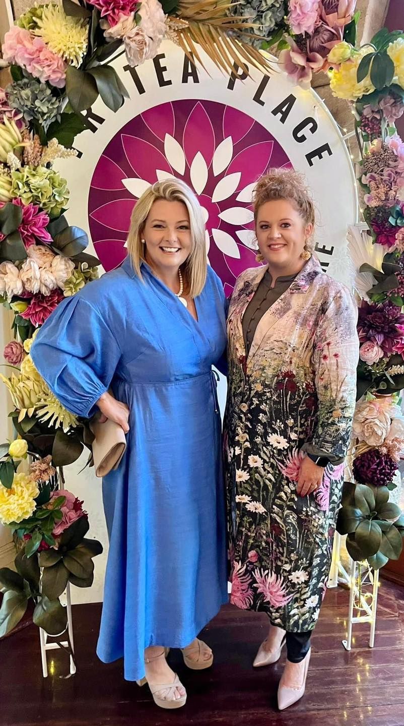 Two women are posing for a picture in front of a wall of flowers.