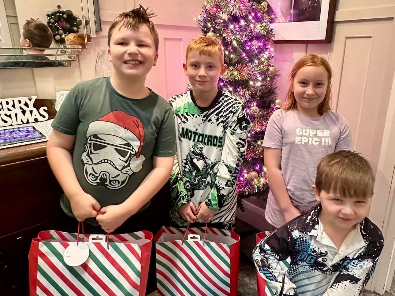 A group of children are holding gift bags in front of a Christmas tree.