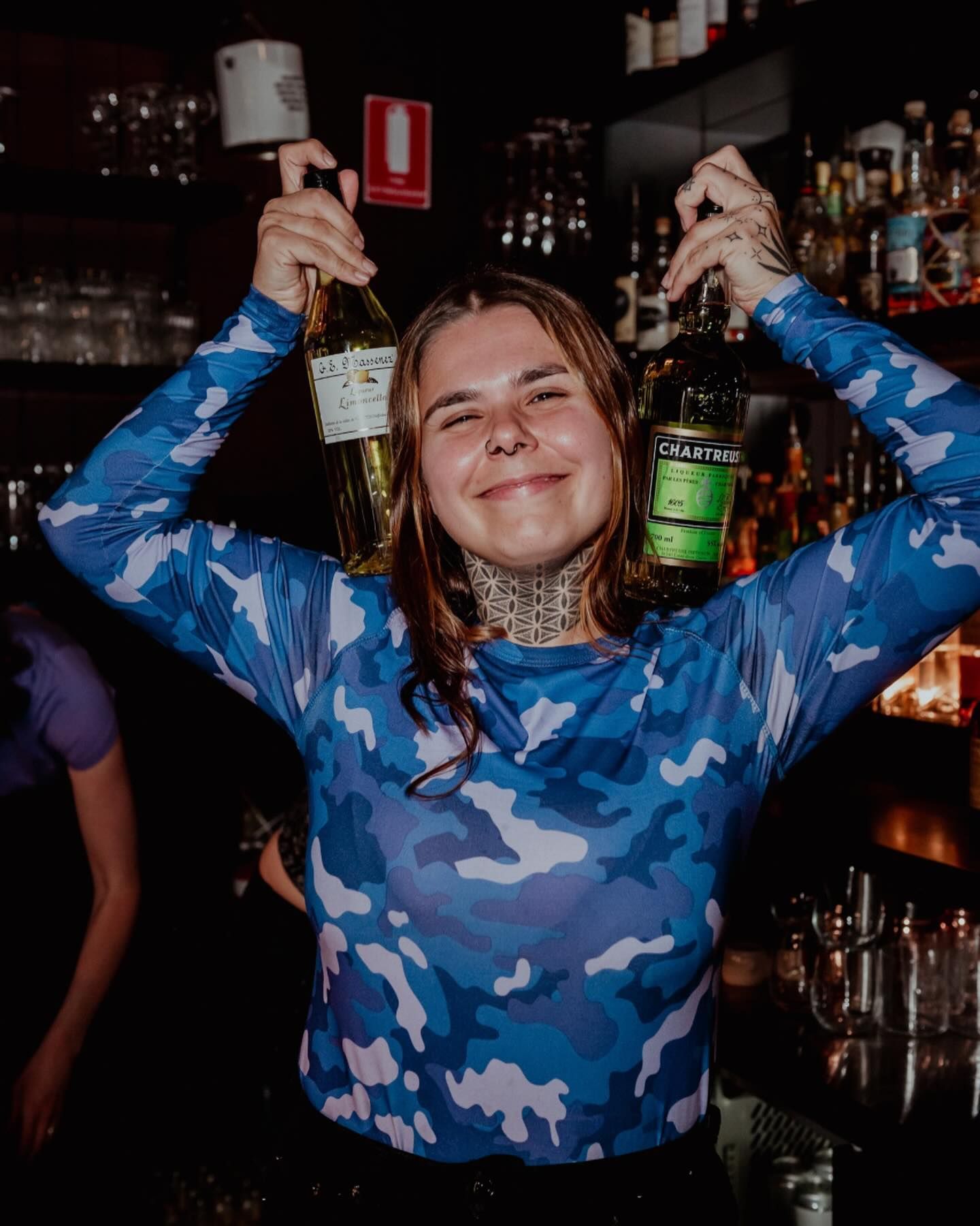 A woman in a blue camouflage shirt is holding a bottle of alcohol over her head.