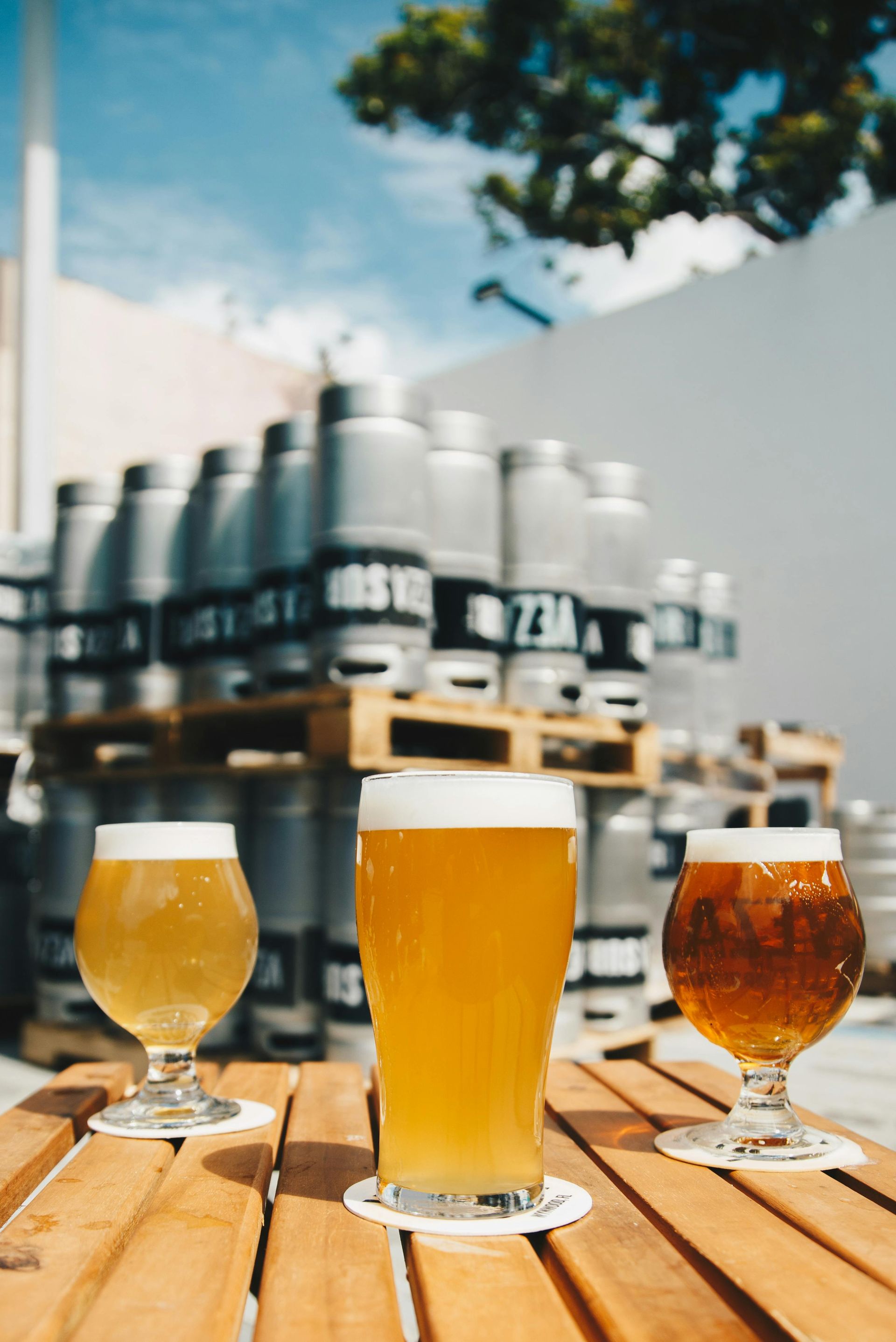 Three beers on wooden table, kegs in background under blue sky.