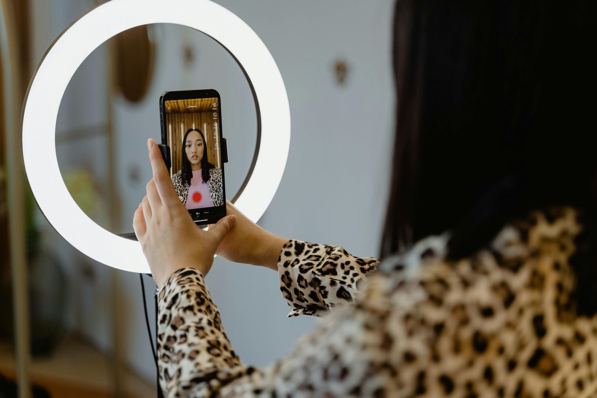 Woman filming a video on her phone, lit by a ring light. She is wearing a leopard print top.