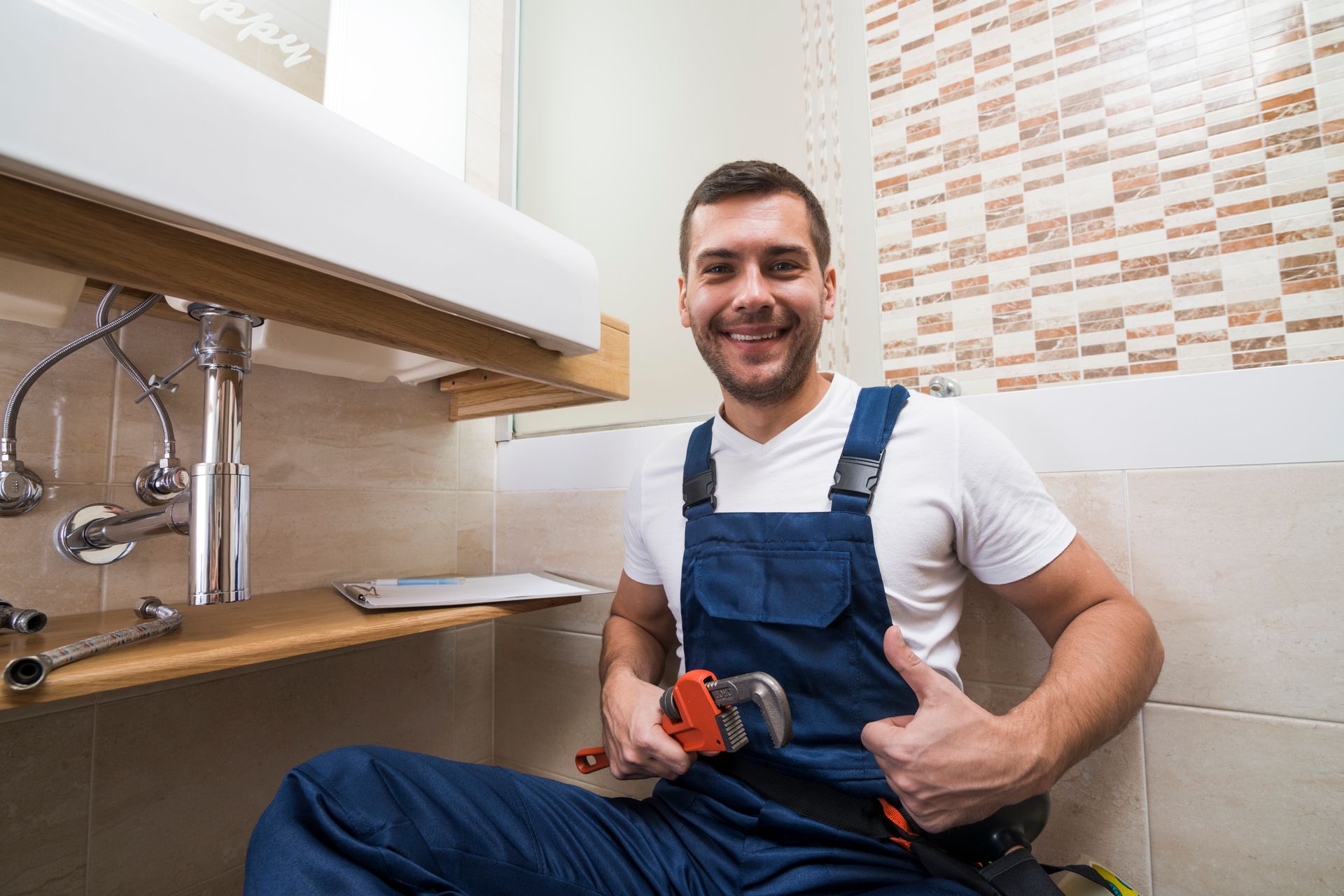 A plumber is sitting under a sink in a bathroom holding a wrench and giving a thumbs up.