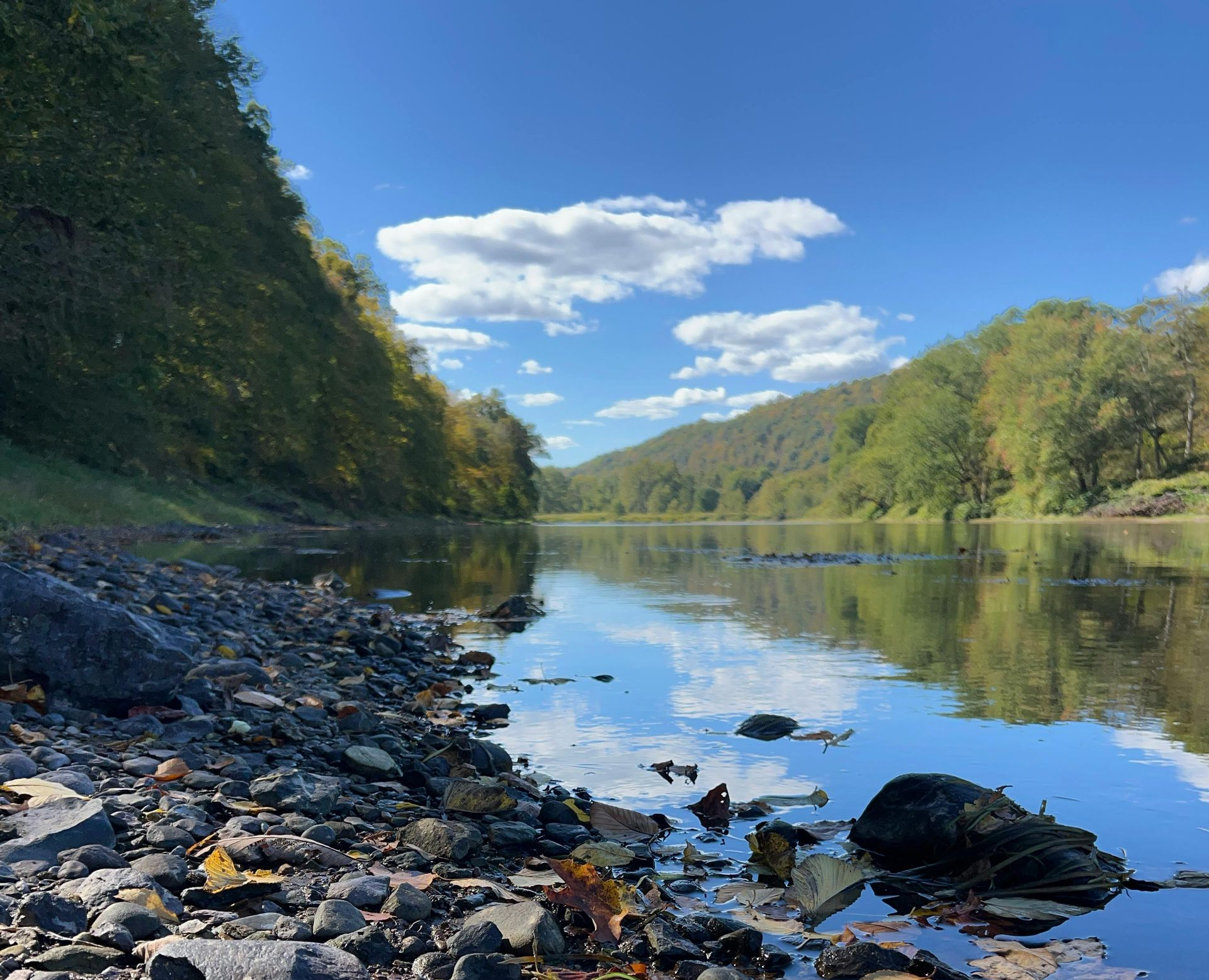 Calm river reflects blue sky and trees; rocky shore in foreground.