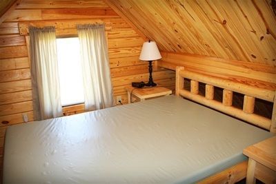 A bedroom in a log cabin with a bed , nightstand , lamp and window.